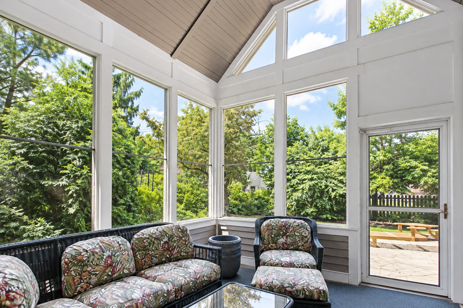 Custom sunroom with a high, peaked ceiling, large windows, patterned wicker seating, and a garden view.
