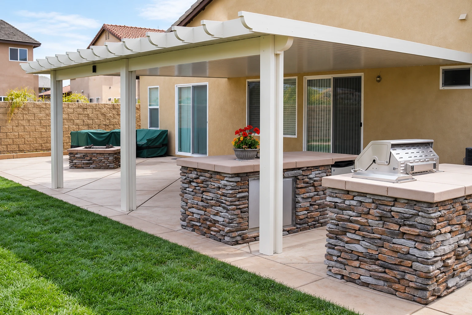 White patio cover shades an outdoor kitchen and paved area beside a tan house.