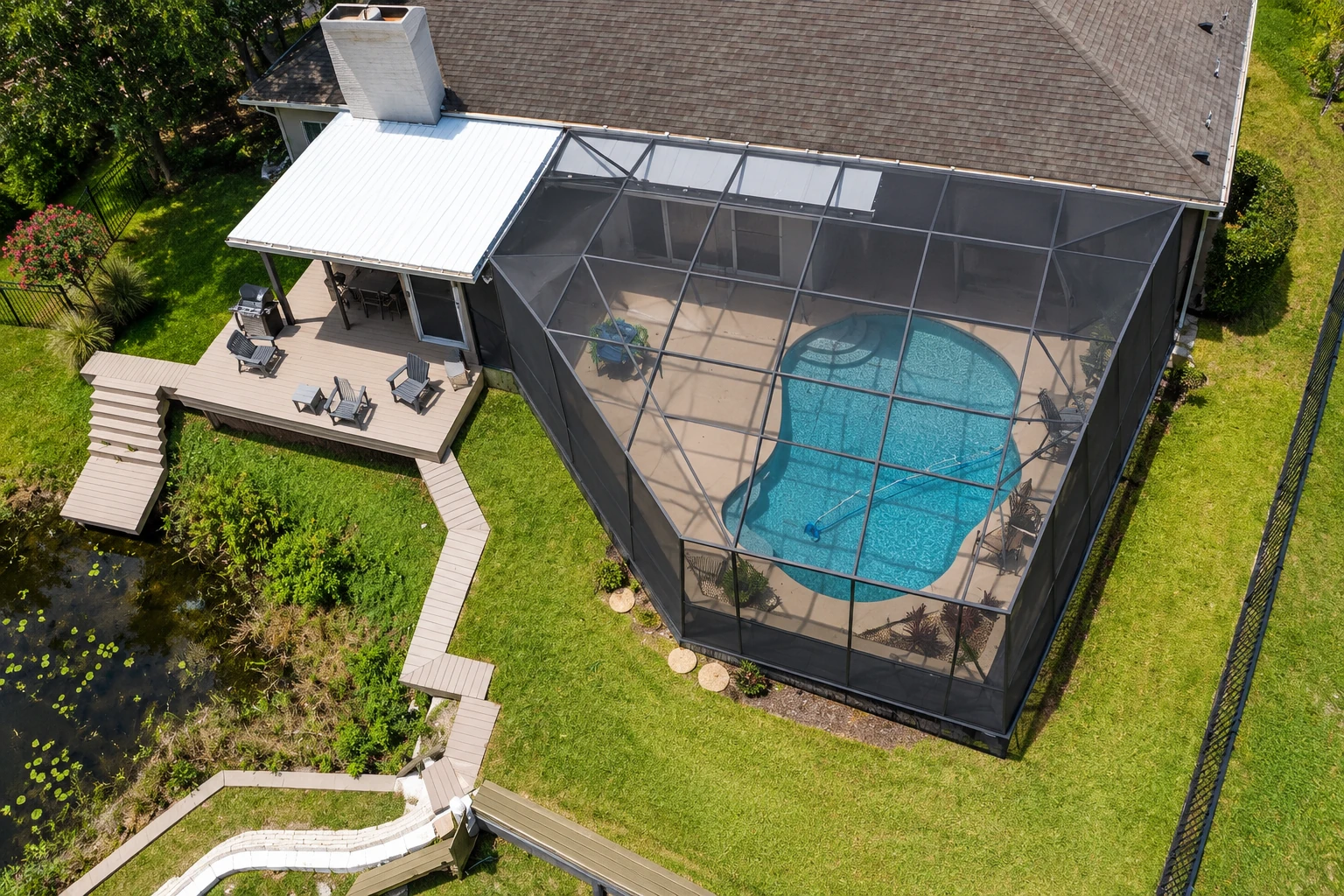 Aerial view of a screened pool enclosure beside a home, with an adjoining deck, lawn, and garden paths.