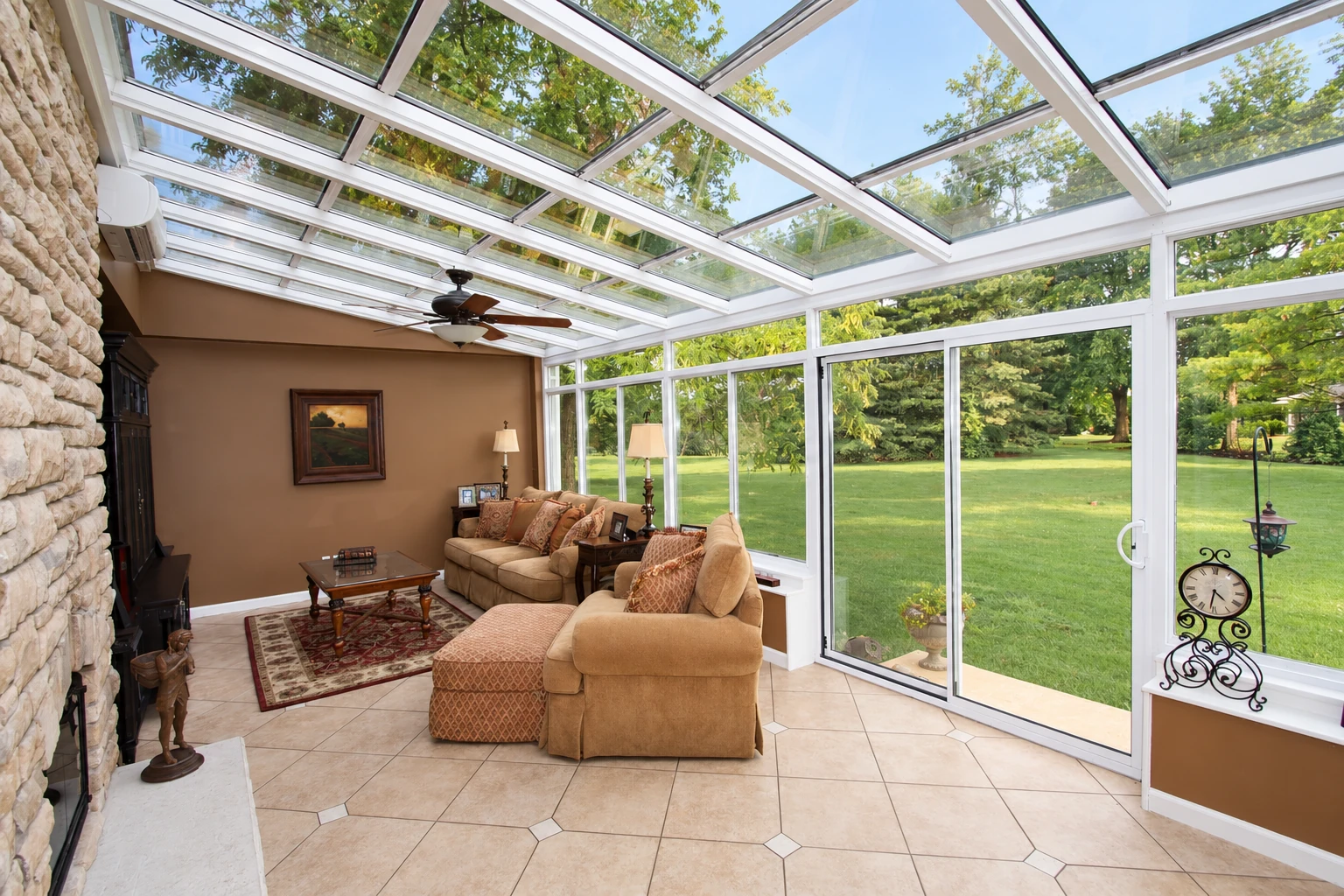 Bright solarium with a glass roof, white-framed windows, tiled floor and tan seating overlooks a lawn.