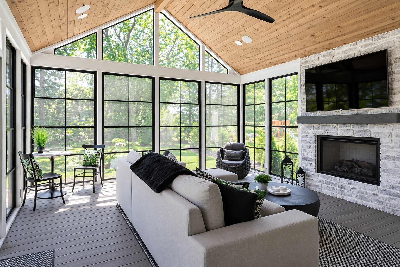 Bright sunroom addition with a peaked wood-paneled ceiling, expansive black-framed windows, seating, and a stone fireplace.