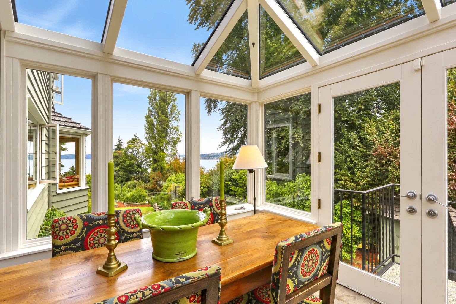 Sunroom dining area with a glass roof, large windows and a wooden table overlooks a leafy garden.